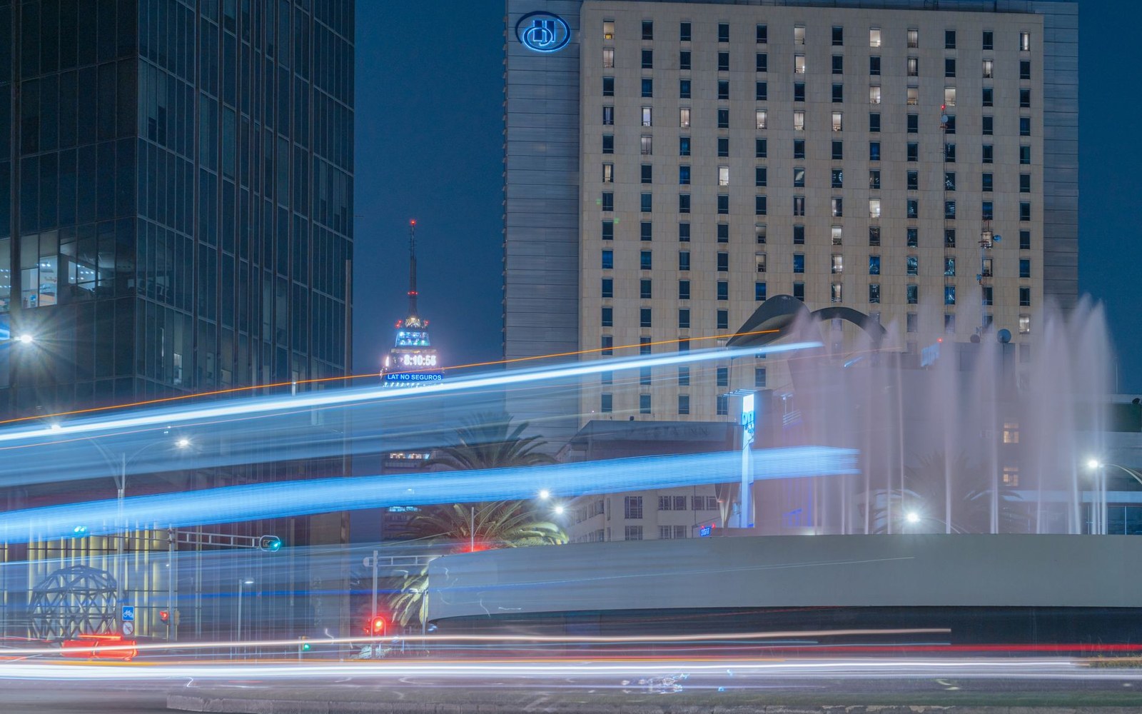 Long-exposure light trails from traffic streak past a fountain and illuminated skyscrapers at night