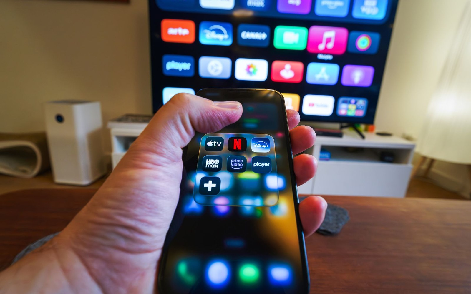 A hand holds a smartphone showing streaming app icons in front of a smart TV home screen
