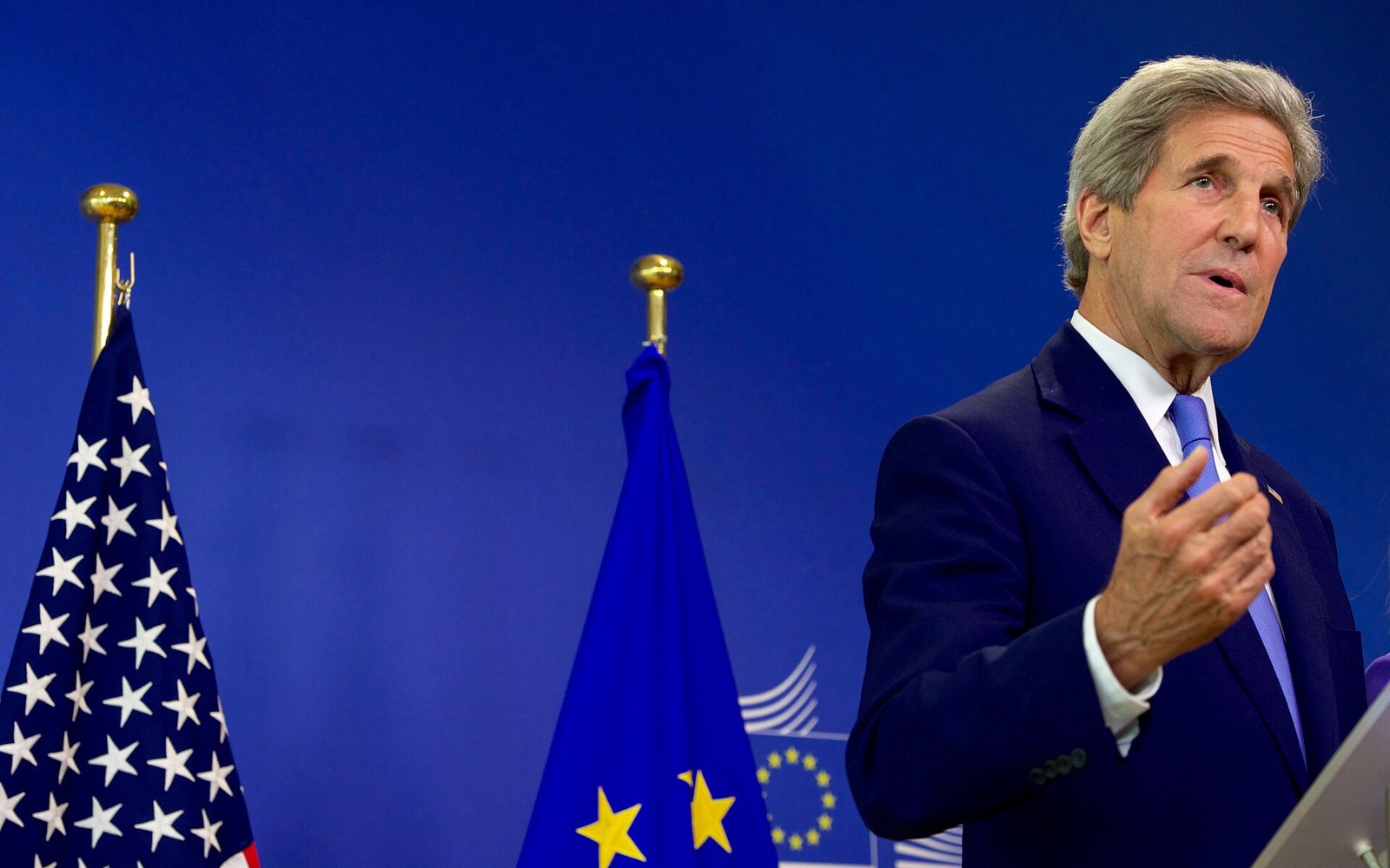 A man in a suit gestures while speaking at a podium beside US and EU flags