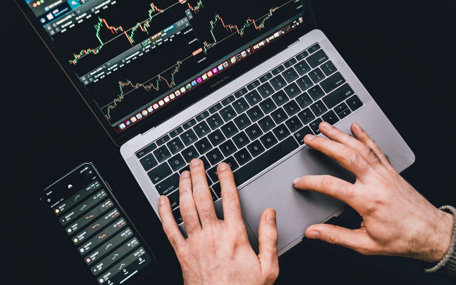 Hands typing on a laptop showing stock charts, with a smartphone displaying trading data nearby